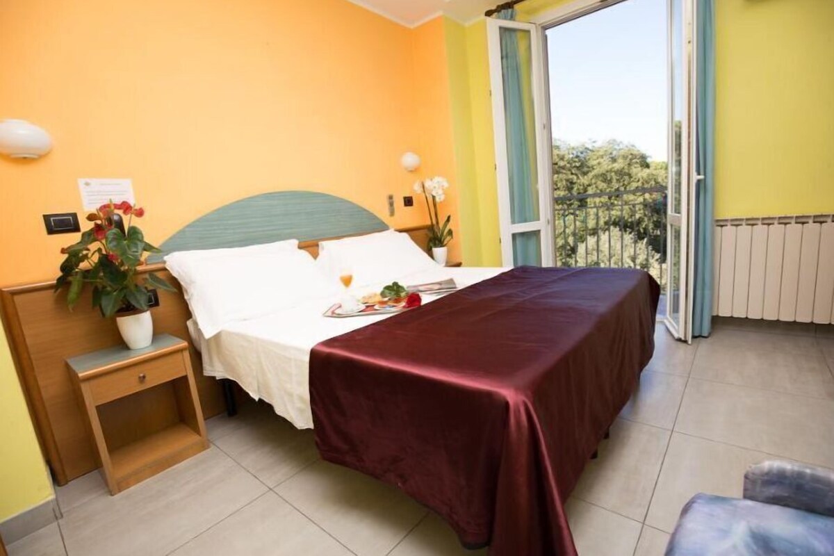 Une chambre à coucher avec un lit, une table de chevet, une plante en pot et une vue sur l’extérieur, visible à travers une porte ouverte.