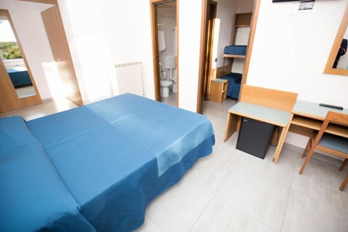 Une chambre comprenant un lit, un bureau, une chaise, un petit réfrigérateur et une salle de bain visible à travers une porte ouverte.