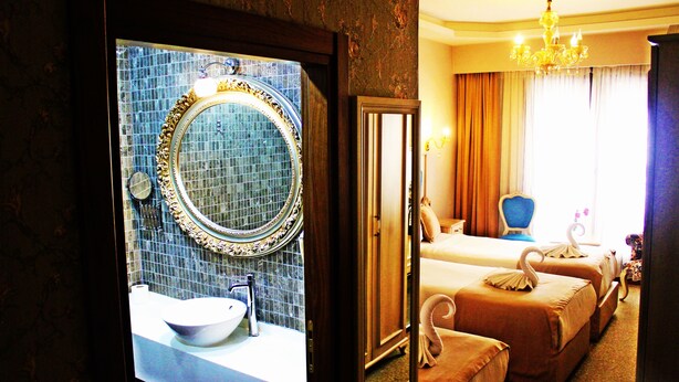 Habitación de hotel con un espejo grande, un espejo redondo, un lavamanos, dos camas con almohadas decoradas con forma de cisne, un candelabro y vista a la ciudad por la ventana.