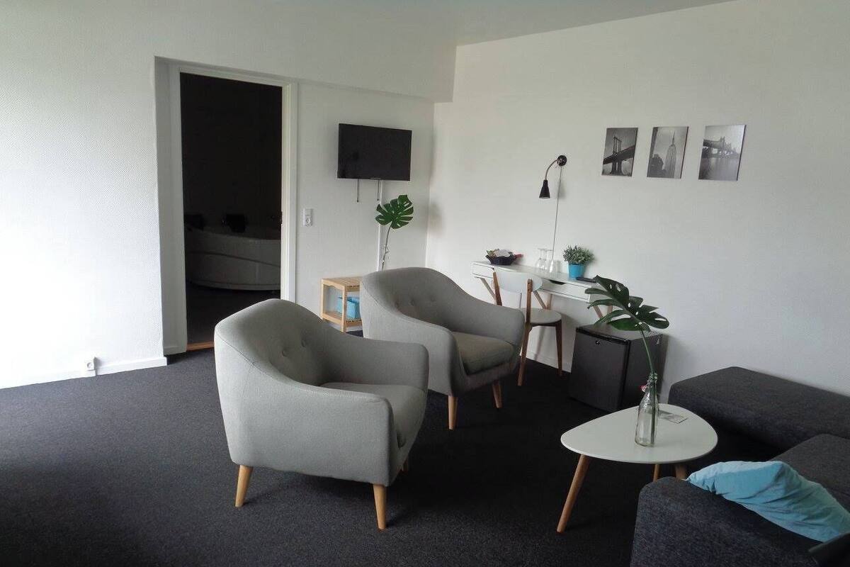 En moderne stue med grå sofaer, lænestole og et lille rundt bord. Der er et TV ophængt på væggen og et par billeder i rammer.