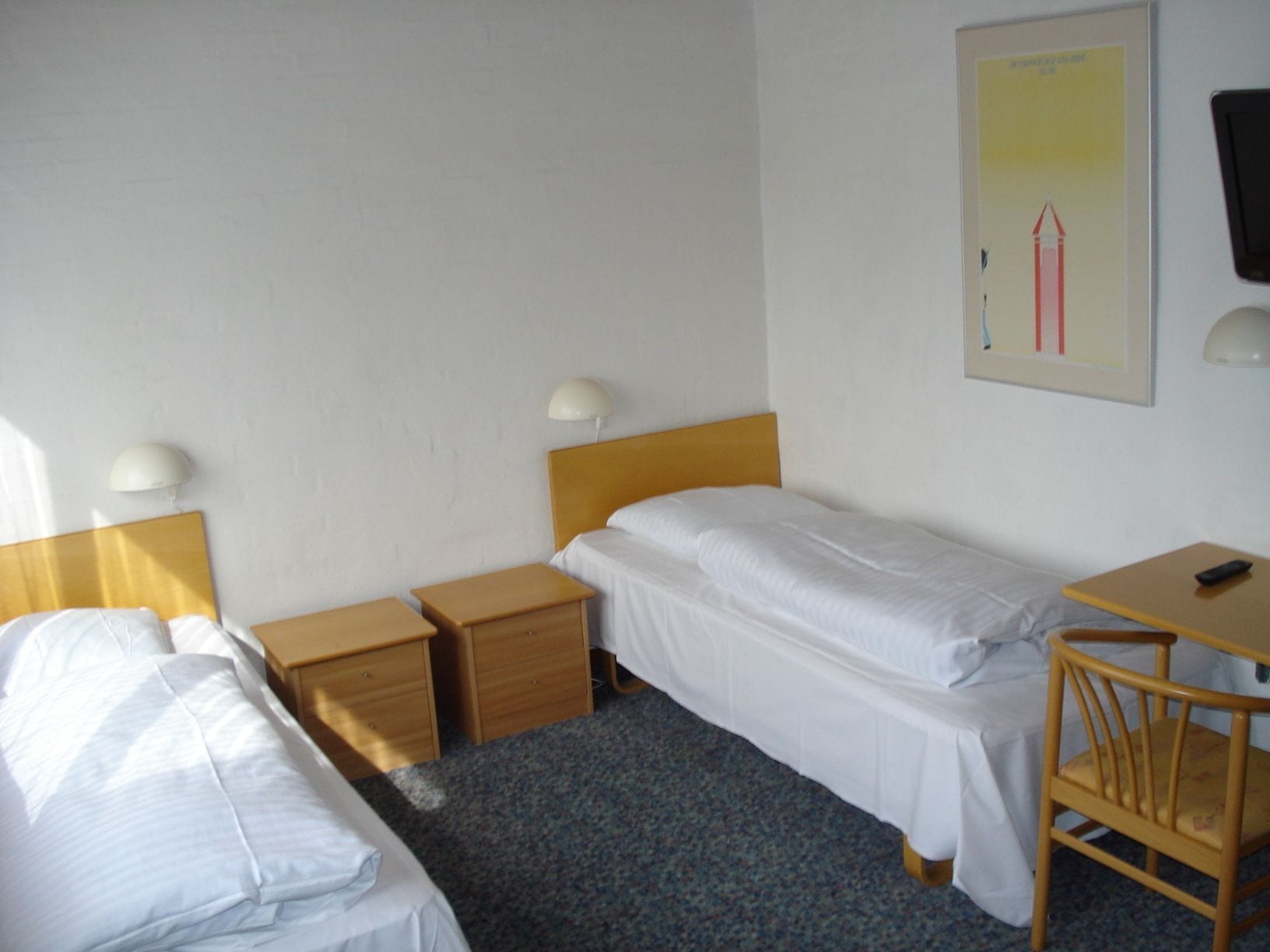 A hotel room with two single beds, a desk with a chair, a nightstand, and a wall-mounted lamp.