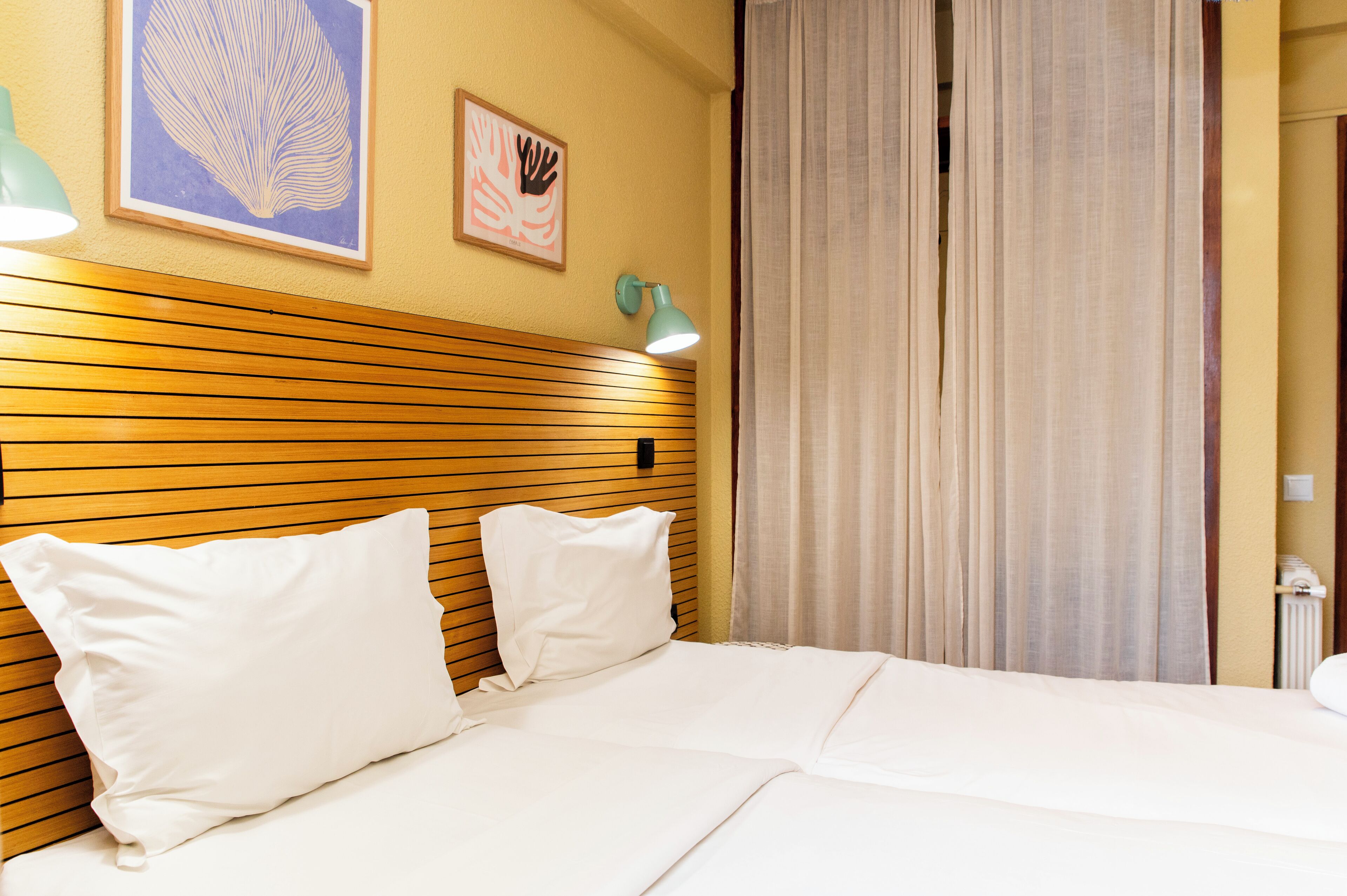 Hotellihuone, jossa on puinen pääty, kaksi valkoista tyynyä, valkoisilla lakanoiden peitetty sänky, turkoosinsininen seinävalaisin ja kaksi kehystettyä taideteosta seinällä.