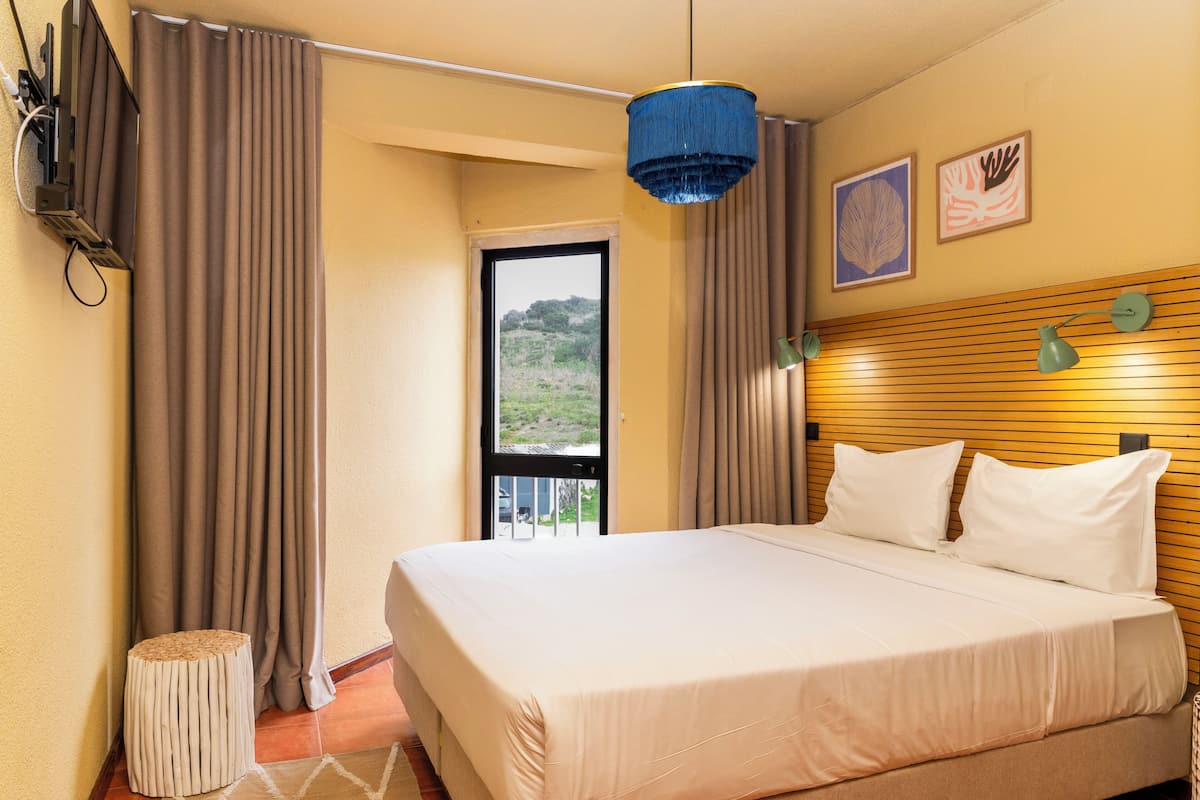 A hotel room with a bed, curtains, a wooden headboard, a ceiling light, and a view of the outdoors.
