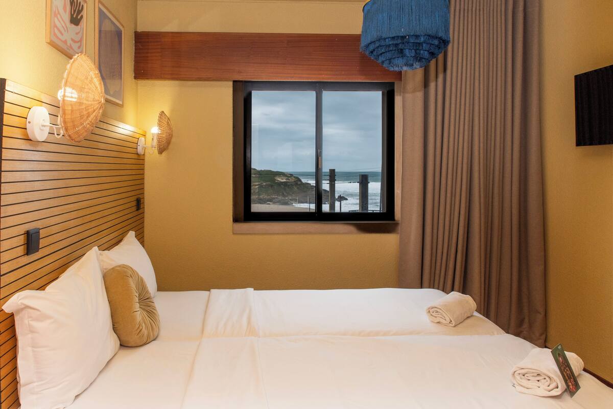 A hotel room with a bed, a window with a view of the sea, a wall-mounted fan, and a television.