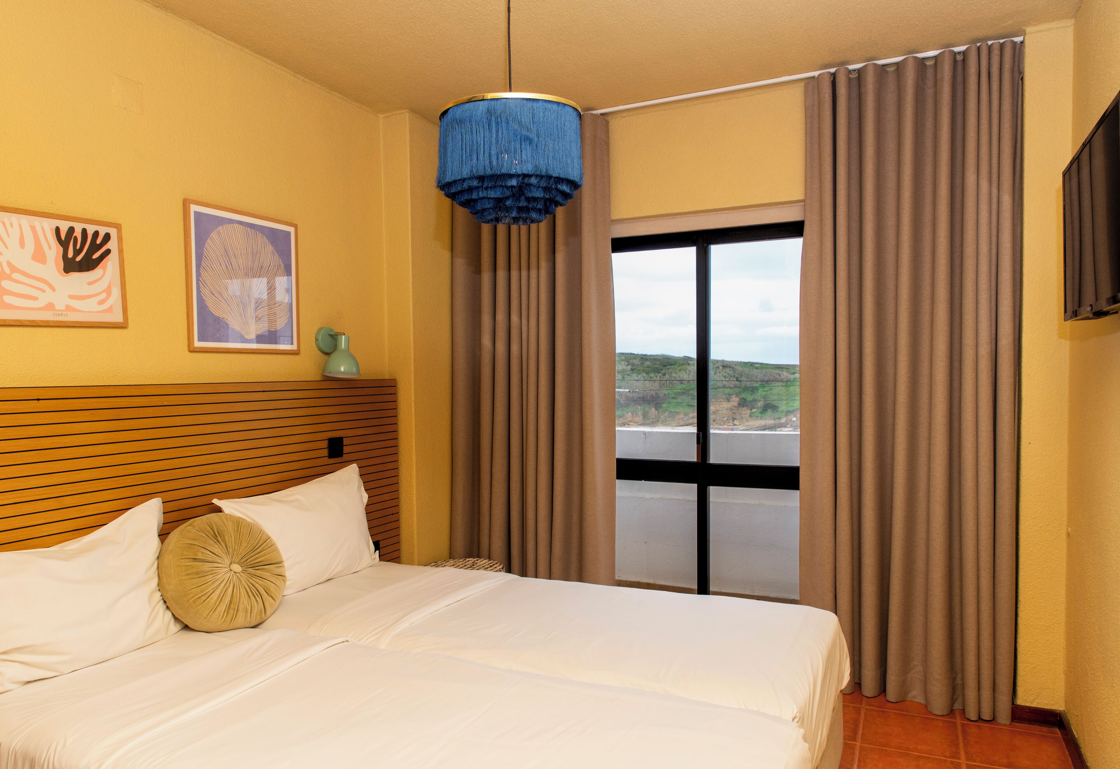 Une chambre d’hôtel avec un lit, un luminaire suspendu bleu, une fenêtre avec des rideaux et deux tableaux encadrés accrochés au mur.