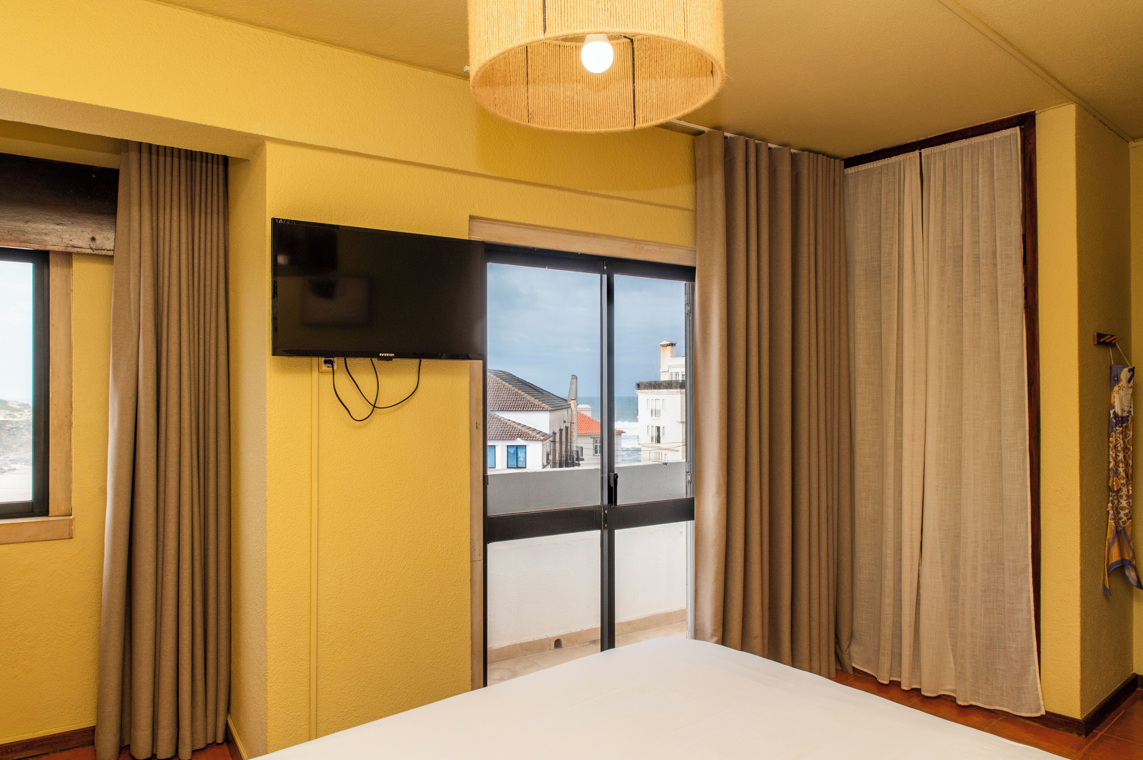 Una habitación de hotel con cama, televisor montado en la pared, ventana con cortinas y vistas a edificios y una masa de agua.