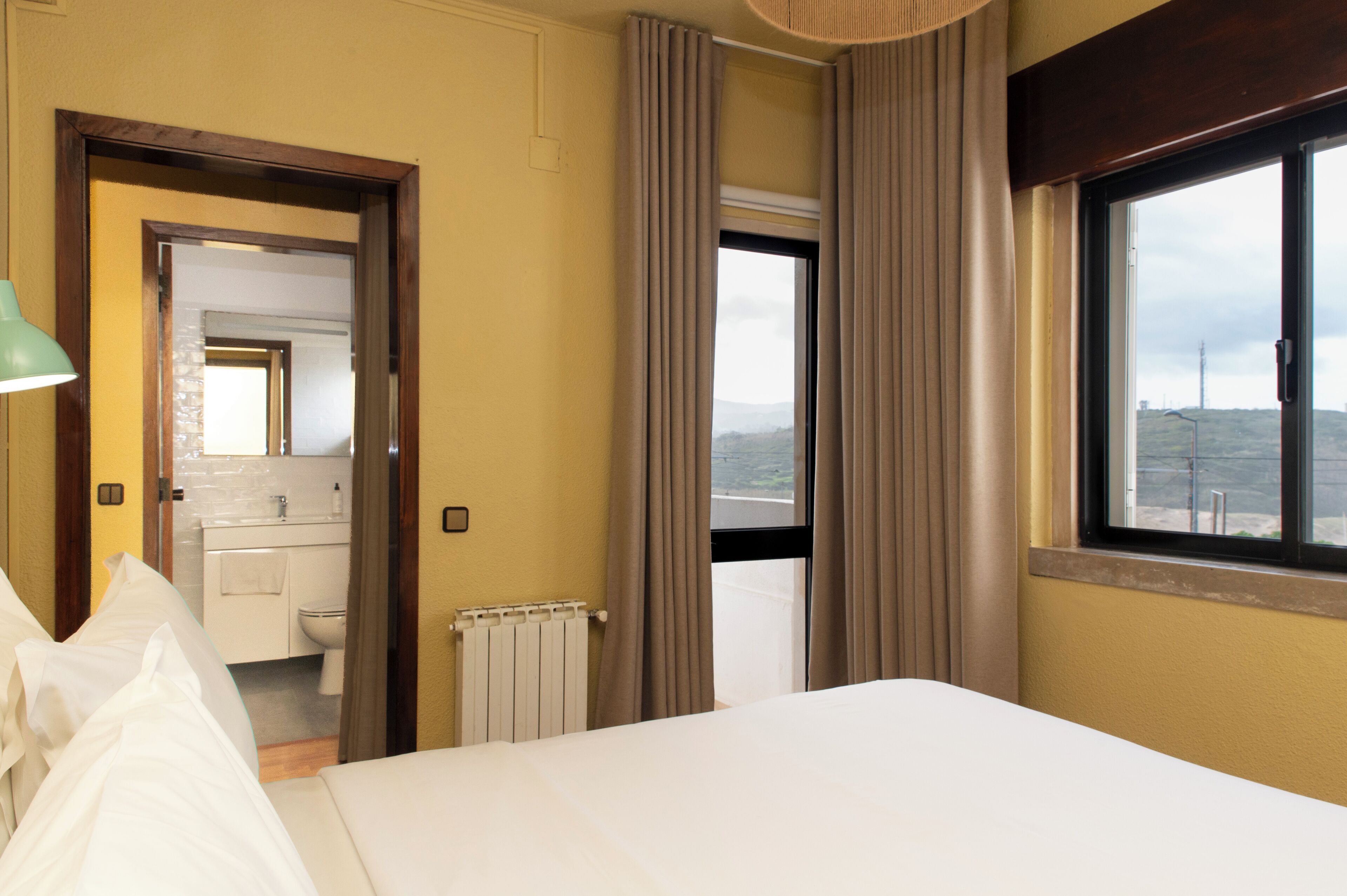 Quarto de hotel com cama, banheiro com vaso sanitário e pia, e janela com vista para a paisagem.