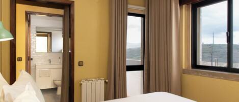 Quarto de hotel com cama, banheiro com vaso sanitário e pia, e janela com vista para a paisagem.