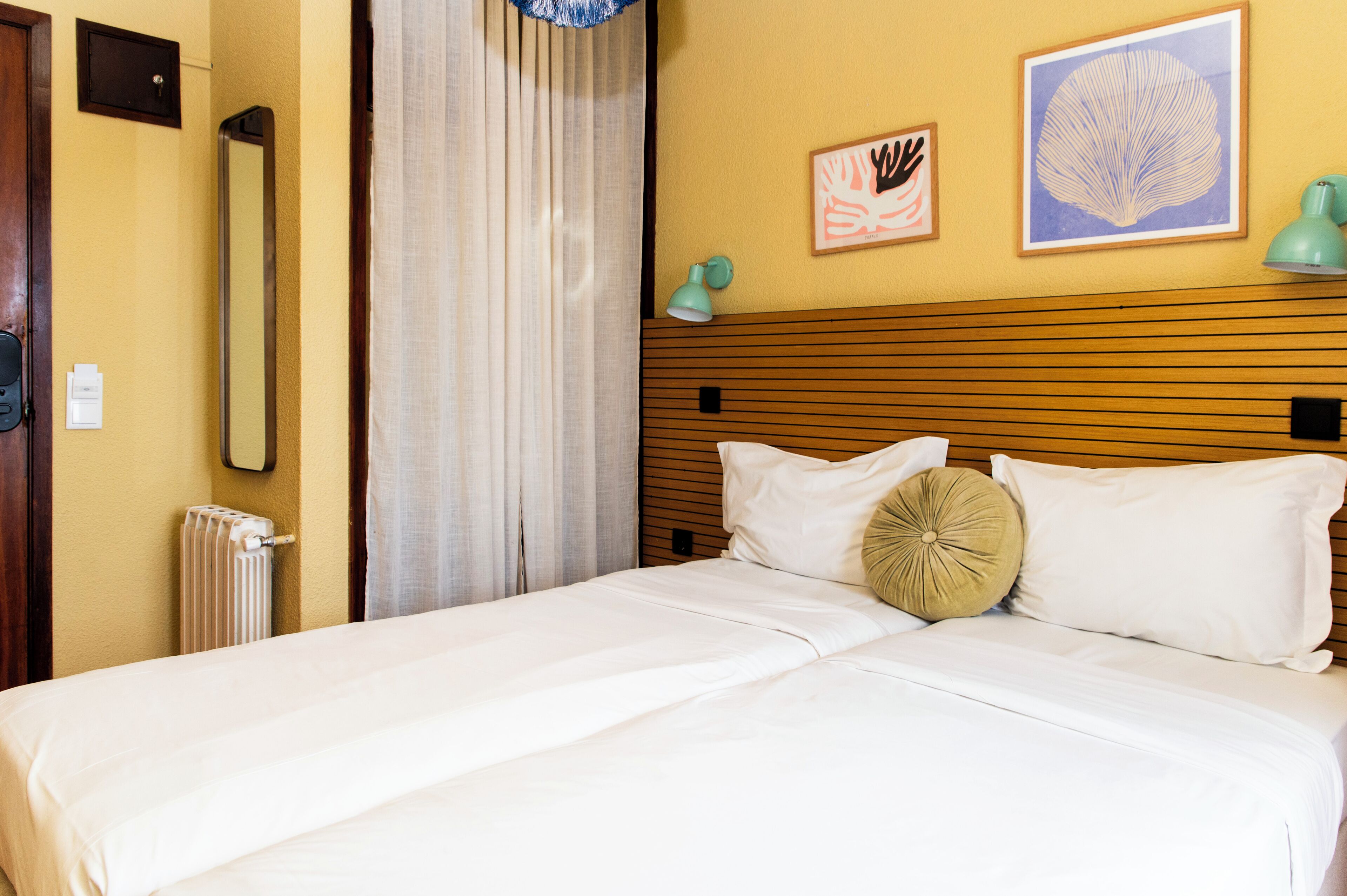Habitación de hotel con cabecera de madera, ropa de cama blanca, un radiador, un espejo y dos cuadros en la pared.