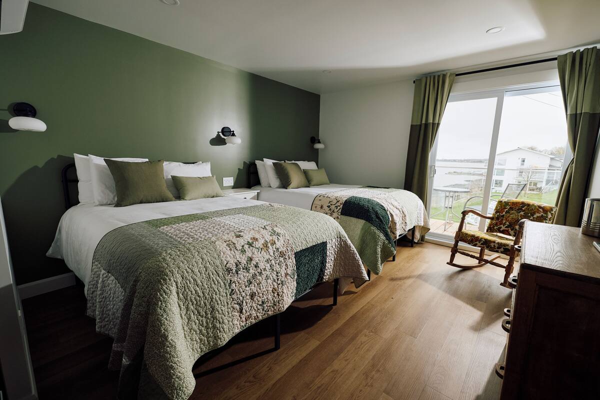 A hotel room with two beds, a rocking chair, a wooden dresser, and a large window with curtains.