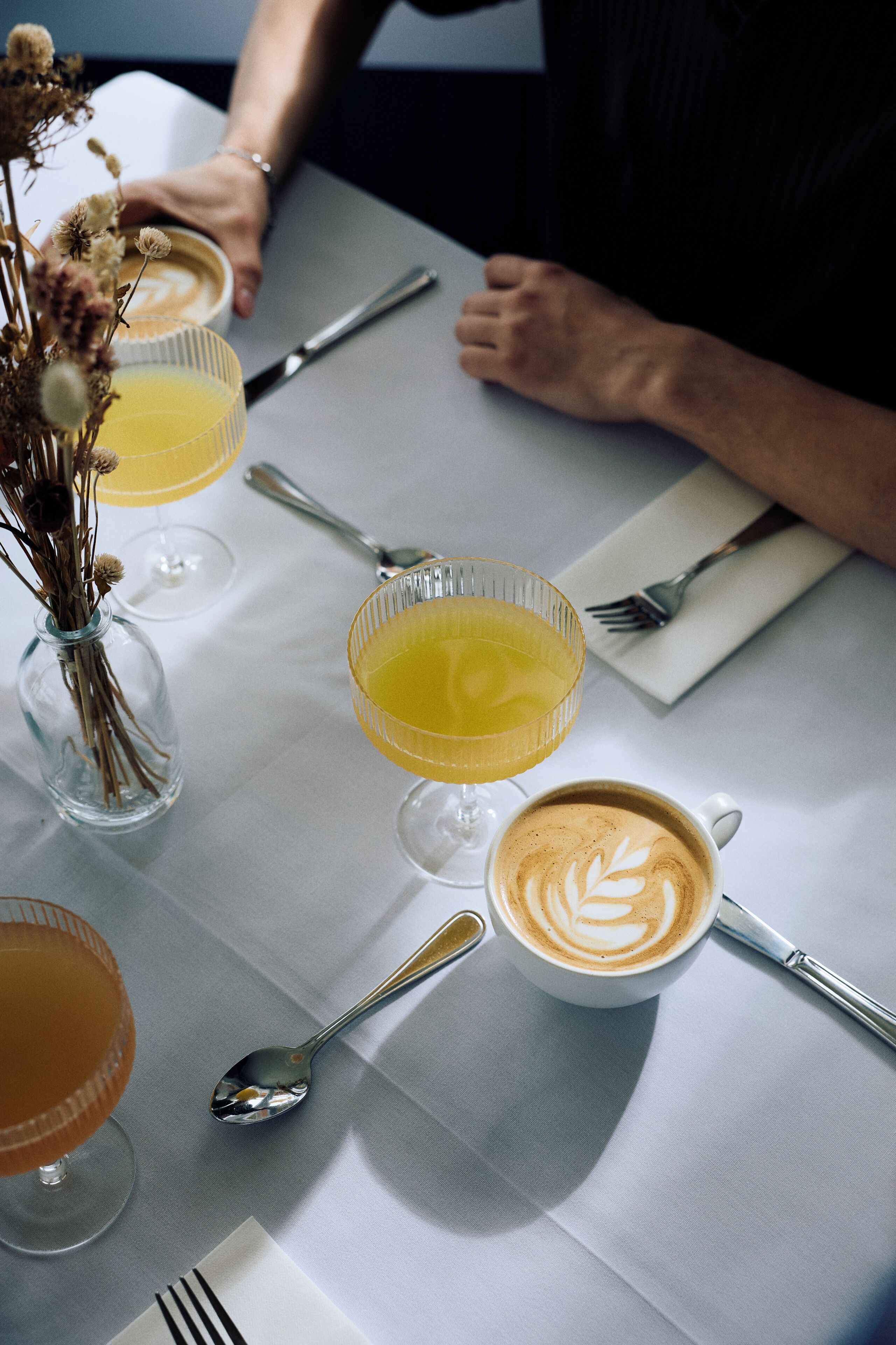 Une table avec un verre de jus d’orange, un verre de café agrémenté d’un motif en latte art, et un autre verre de jus d’orange décoré d’une composition florale.