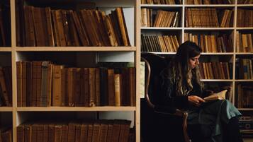 A person seated in a chair reading a book in a room with bookshelves filled with books.