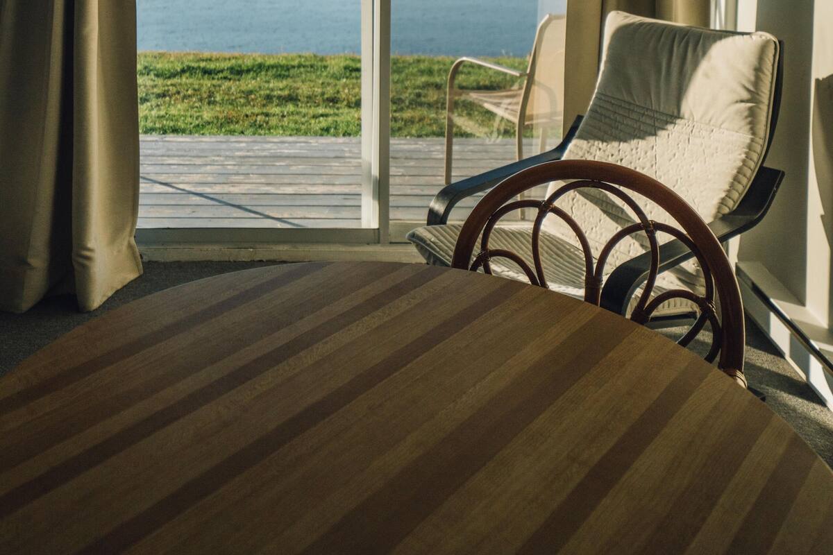 A room with a large window offering a view of the sea. There is a bed with a striped headboard, a chair, and a hanging light fixture.