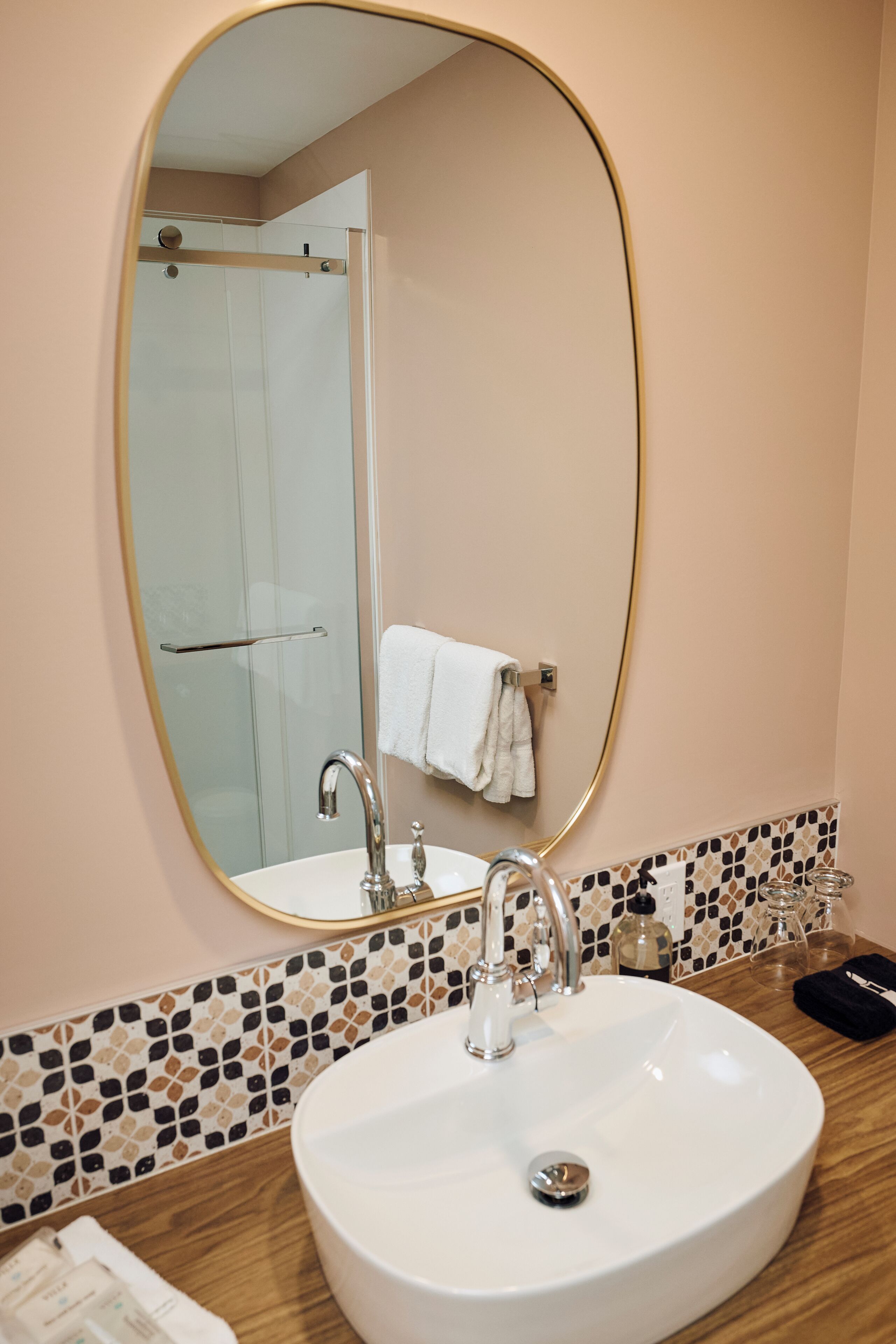 Un baño con un espejo ovalado grande, un lavamanos blanco, una ducha y un salpicadero de azulejos.