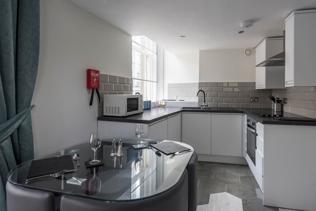 A modern kitchen with white cabinets, a microwave, and a sink. There is a dining table with a glass top and a few wine glasses.