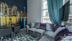Un salon moderne avec une fresque représentant un paysage urbain, un canapé gris agrémenté de coussins, une table à manger et une grande fenêtre avec des rideaux.