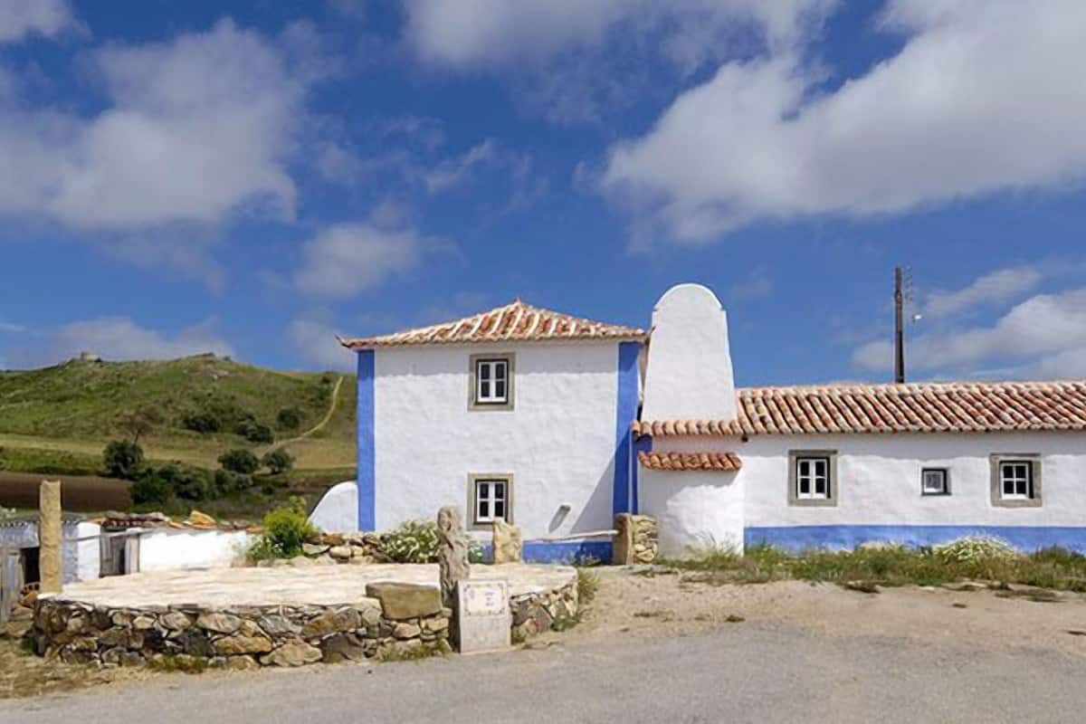 A rural house with a white exterior and blue trim, surrounded by a stone wall and a stone pathway.