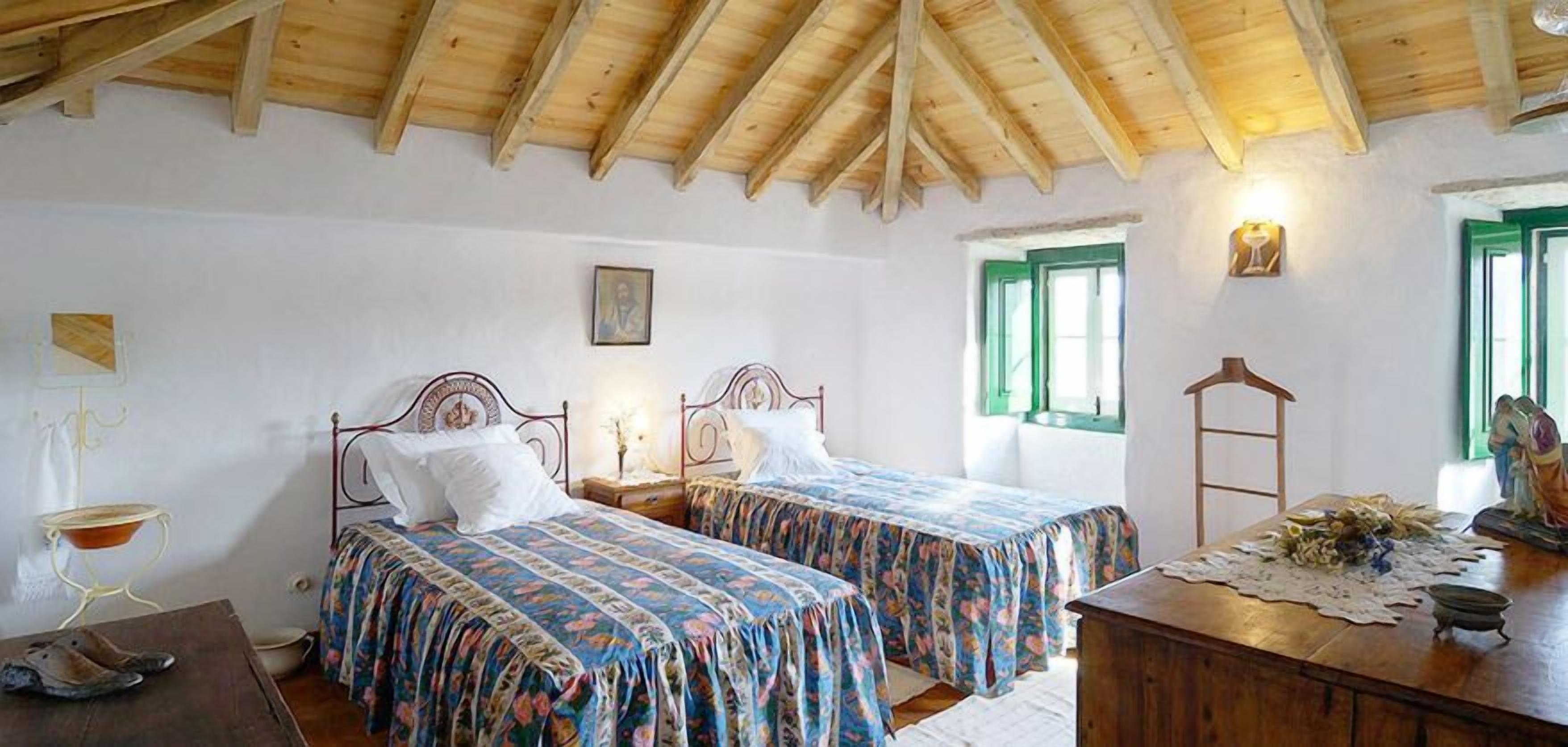 Ein Schlafzimmer mit zwei Betten, Holzbalkendecke, einem kleinen Tisch und einem Fenster mit grünen Rollläden.