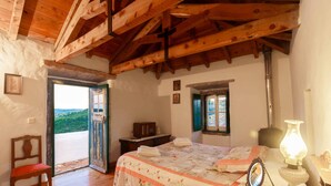A bedroom with a wooden ceiling, a bed with a floral quilt, a bedside table with a lamp, and a view through an open door. - Aldeia da Mata Pequena (Mafra)