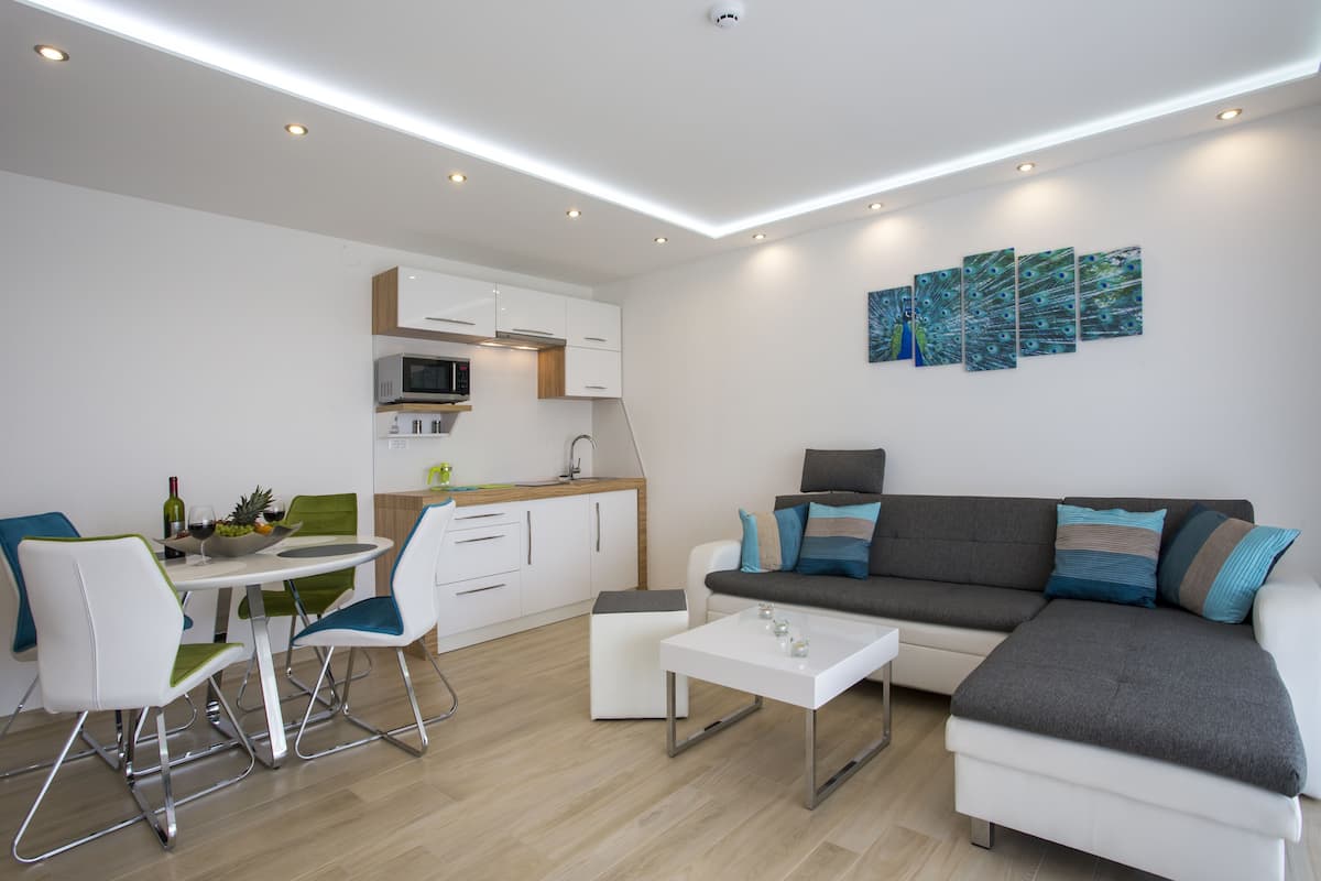 Una sala de estar moderna con un sofá gris, una mesa de centro blanca y sillas verdes. Hay una zona de cocina con armarios blancos y un microondas.
