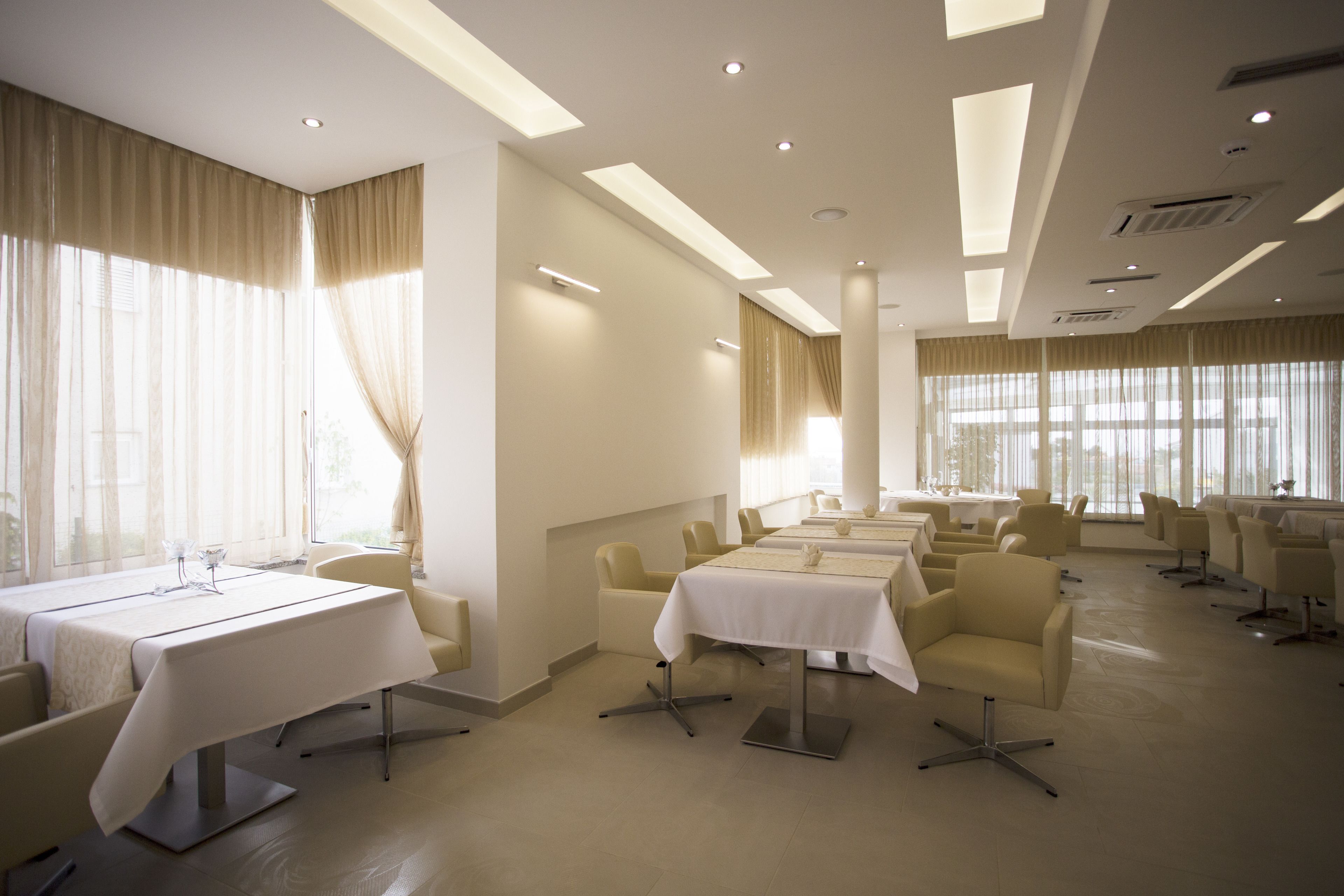 Un salón de conferencias de hotel con varias mesas y sillas, ventanas amplias con cortinas e iluminación empotrada en el techo.