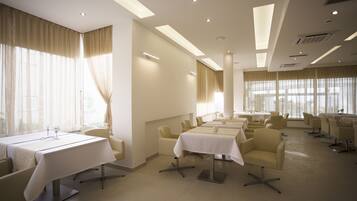 Un salón de conferencias de hotel con varias mesas y sillas, ventanas amplias con cortinas e iluminación empotrada en el techo.