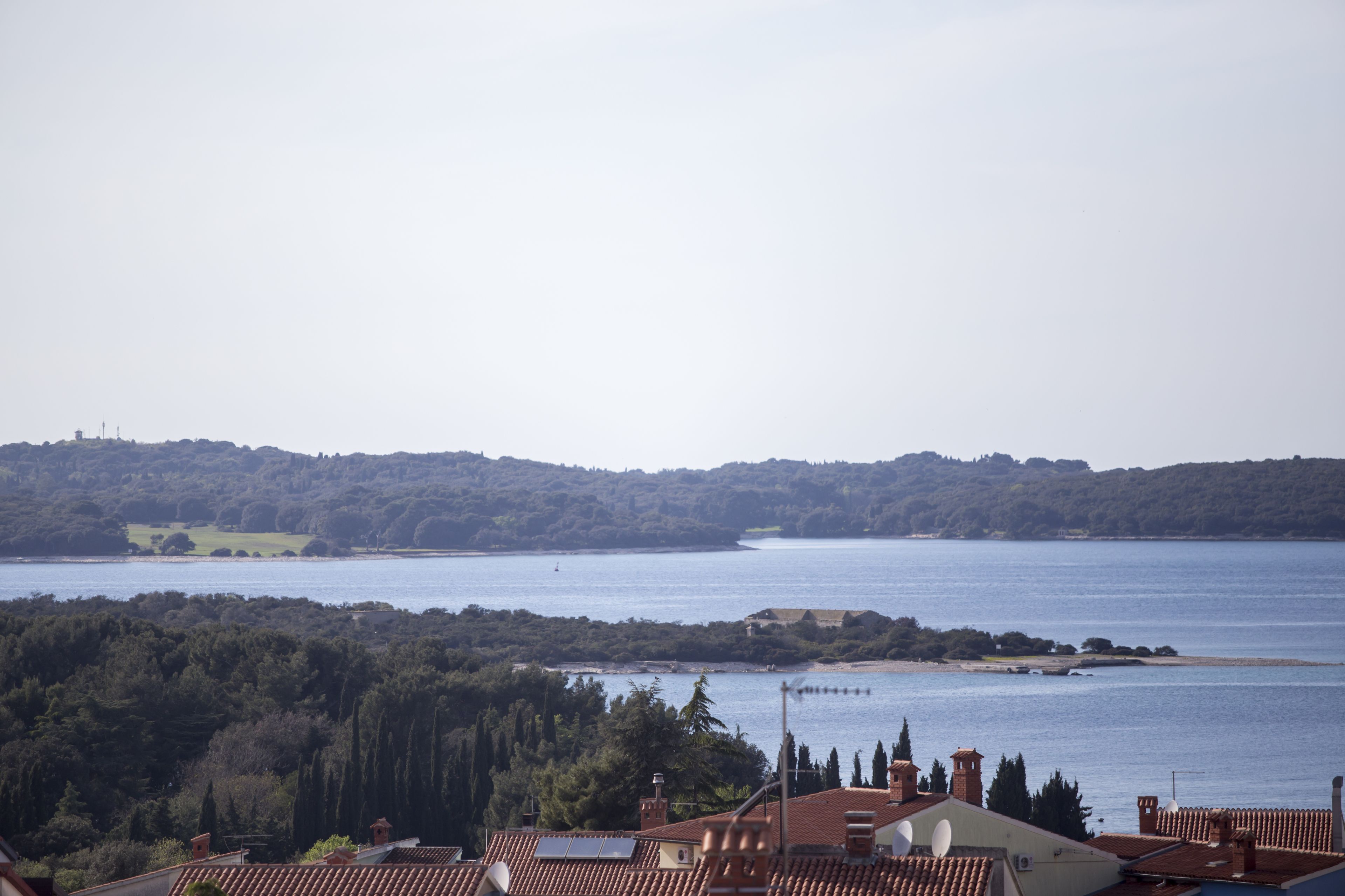 A coastal view with terracotta rooftops, a body of water, and a hilly landscape in the distance.