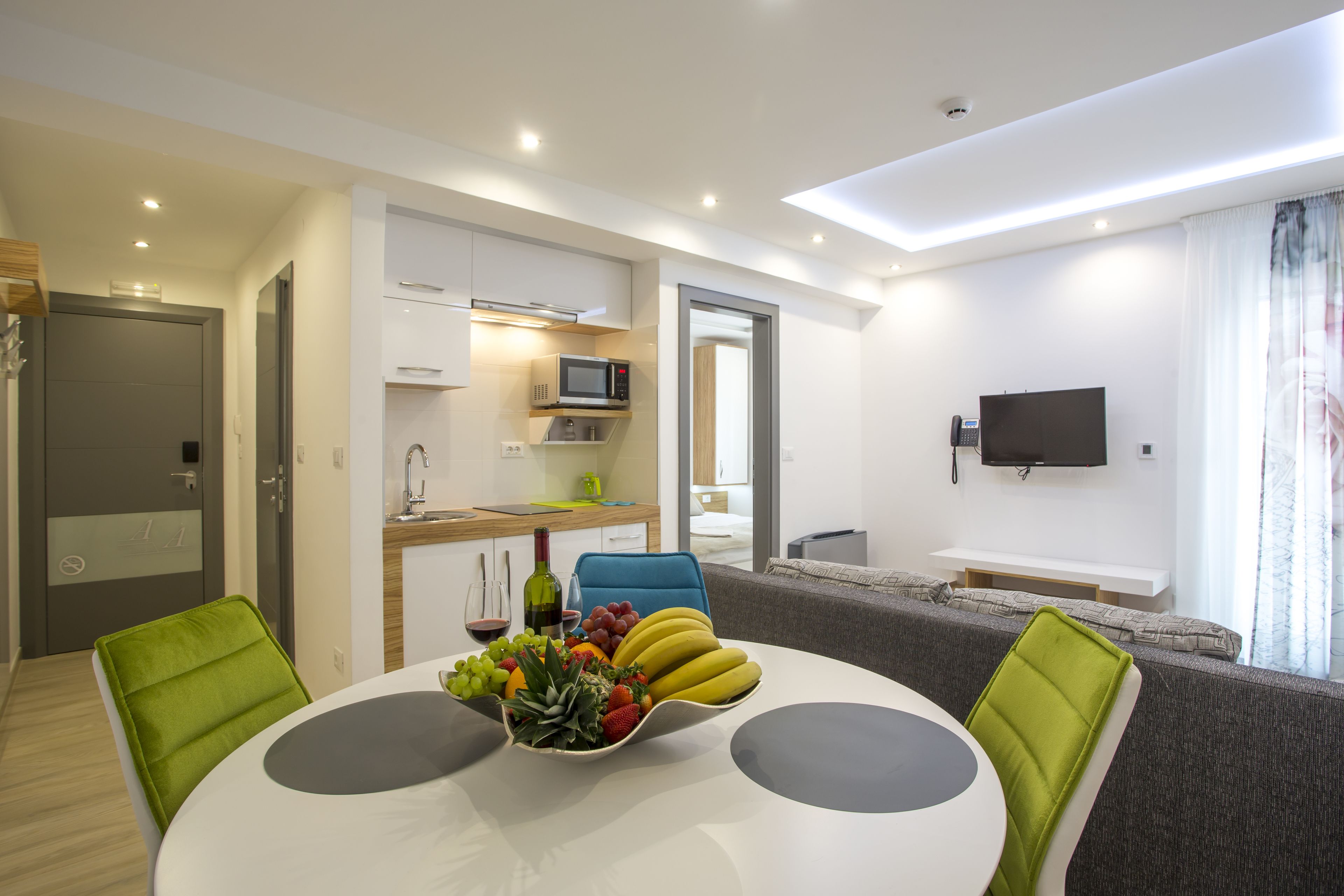 Una sala de estar moderna con una mesa de comedor, sillas verdes, un sofá, un televisor y una zona de cocina con un microondas.
