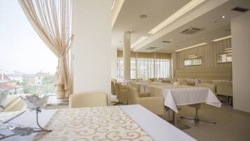 Un comedor de hotel con mesas preparadas para el servicio, ventanas grandes con cortinas y vistas a la ciudad.