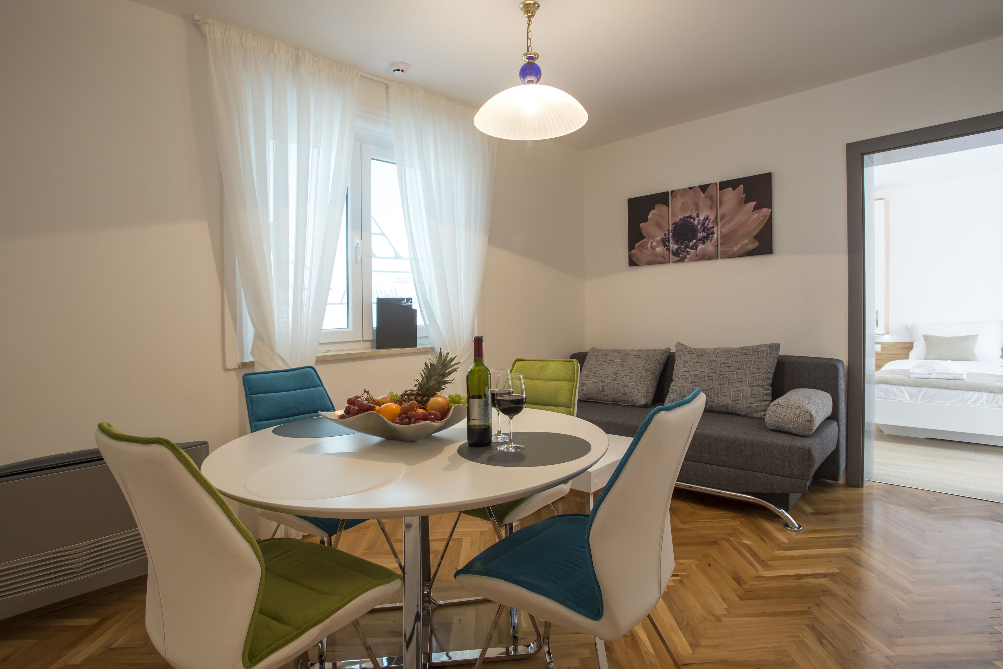 A modern living room with a dining area, featuring a round table with chairs, a sofa, and a large window with curtains.