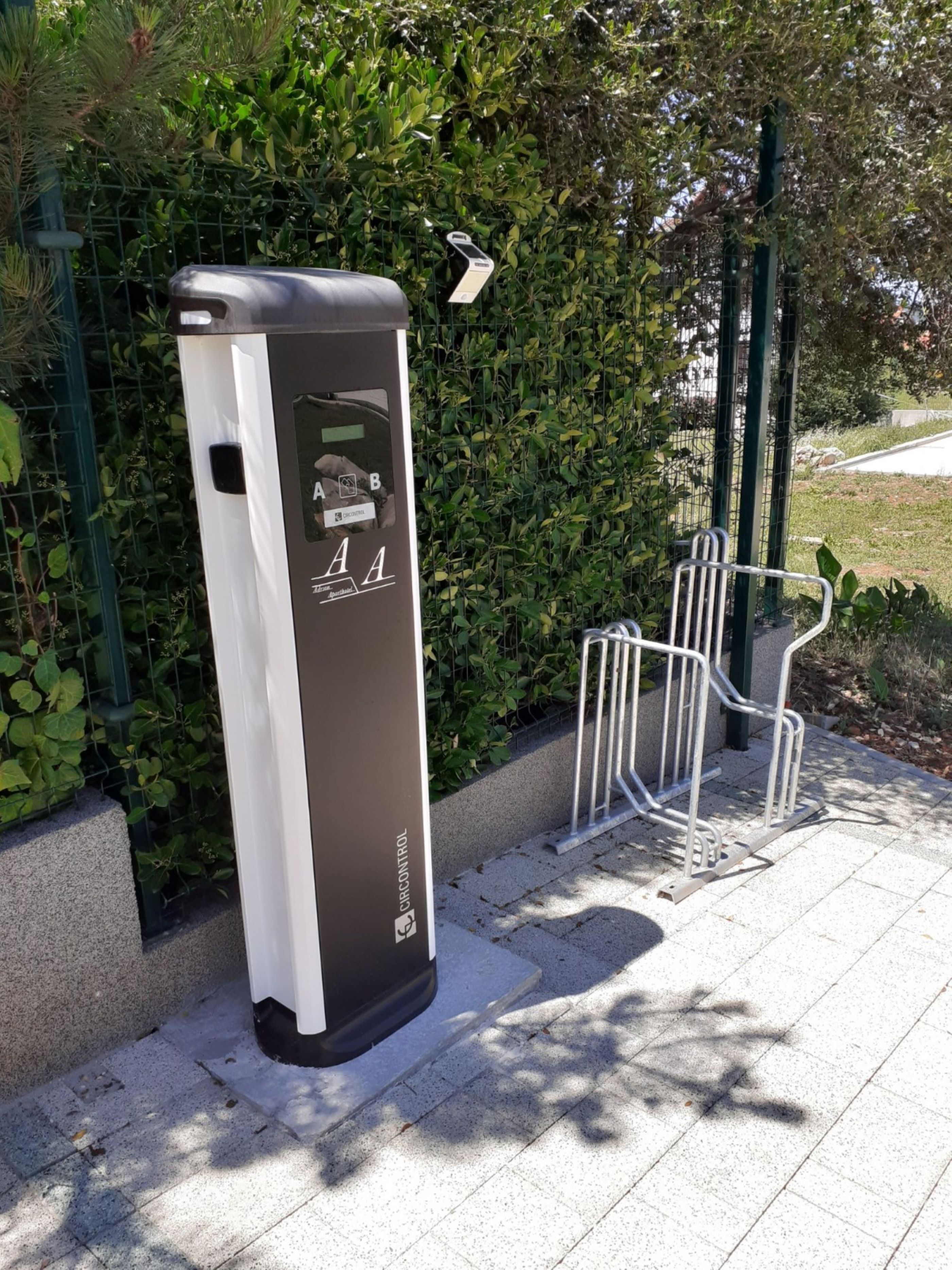 En modern, rektangulär laddstation för elfordon med en digital display och laddsymboler, placerad på en asfalterad trottoar med ett metallstaket och grönska i bakgrunden.