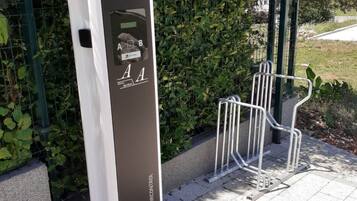 En modern, rektangulär laddstation för elfordon med en digital display och laddsymboler, placerad på en asfalterad trottoar med ett metallstaket och grönska i bakgrunden.