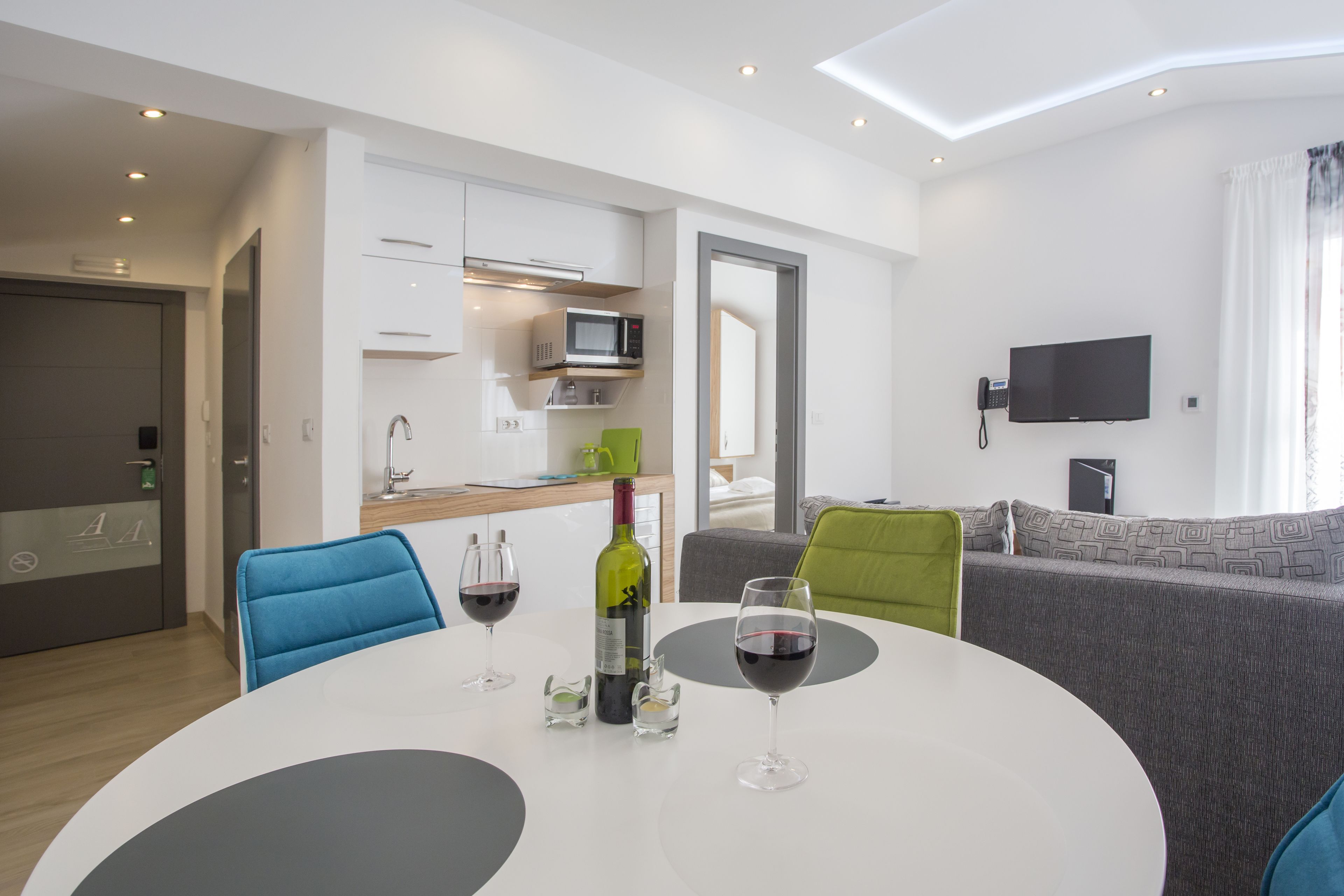 Un salón moderno con una mesa redonda blanca, dos sillas azules, un sofá gris y un televisor de pantalla plana montado en la pared.