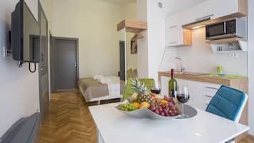 Una habitación de hotel moderna con cama, una mesa de comedor con fruta y vino, una zona de cocina y un televisor montado en la pared.