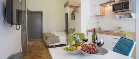 Una habitación de hotel moderna con cama, una mesa de comedor con fruta y vino, una zona de cocina y un televisor montado en la pared.