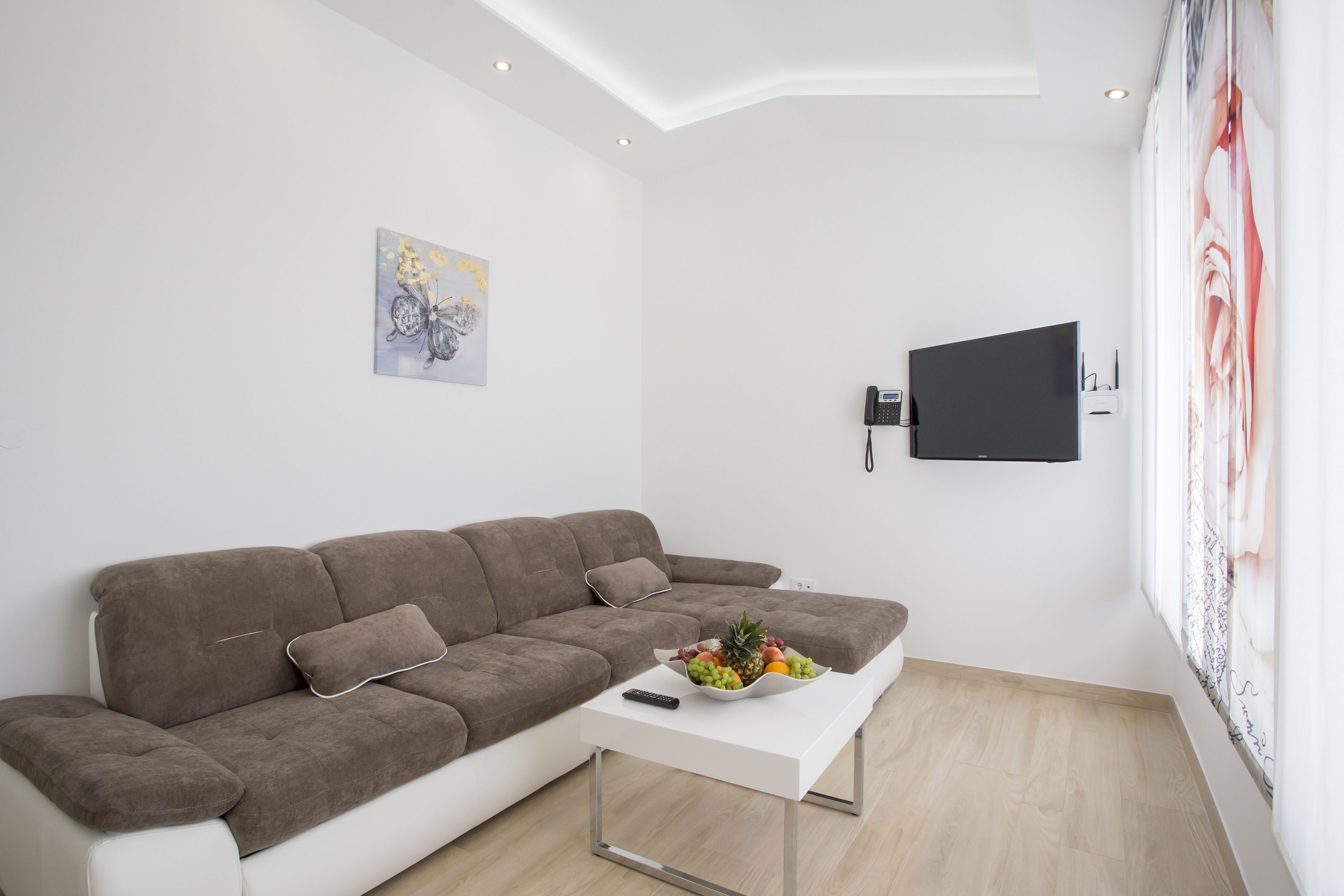 A modern living room with a brown sofa, a white coffee table with fruit, a mounted flat-screen TV, and a painting on the wall.