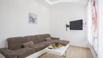 A modern living room with a brown sofa, a white coffee table with fruit, a mounted flat-screen TV, and a painting on the wall.