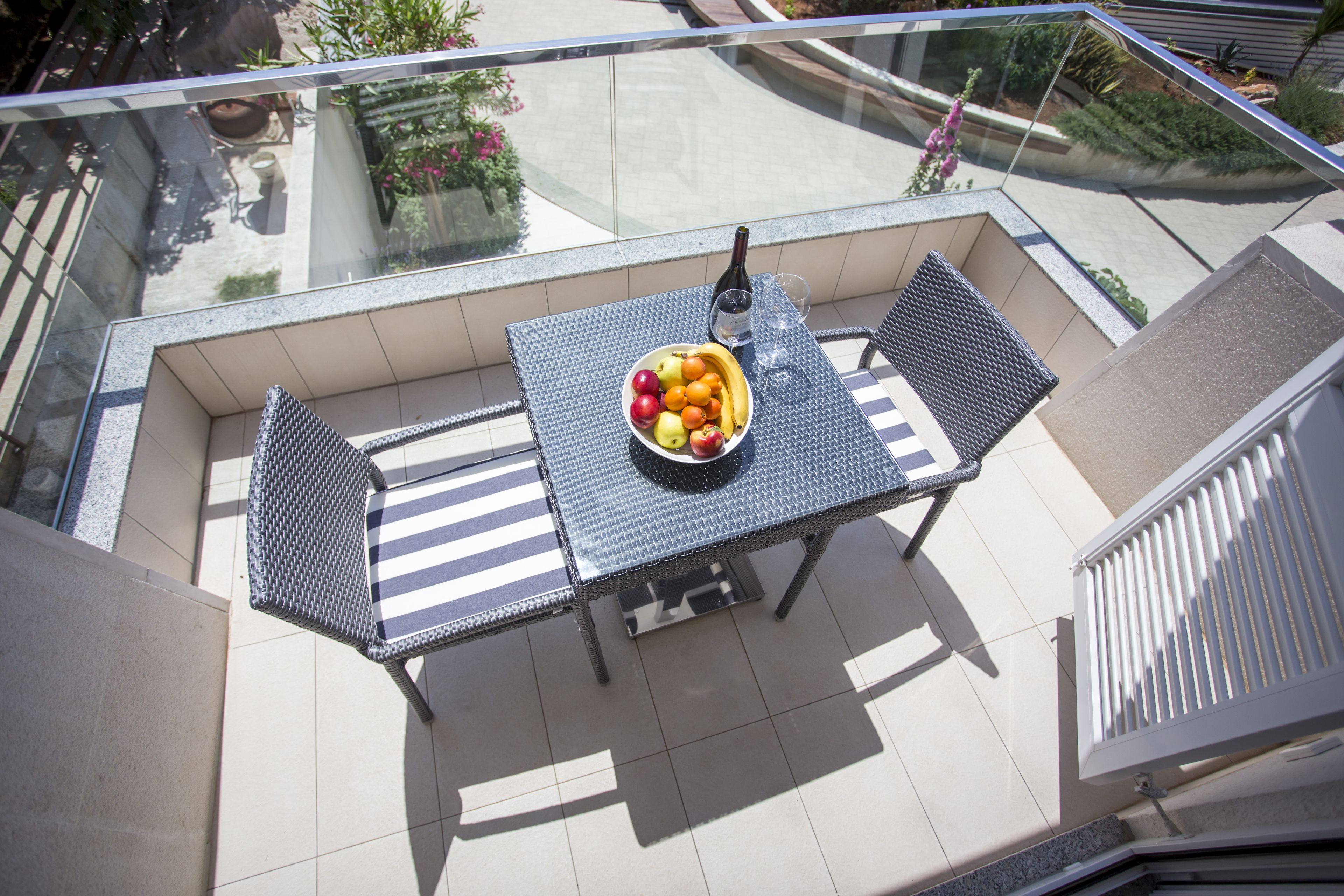 A balcony with a table set for two, featuring a bowl of fruit and a bottle of wine.