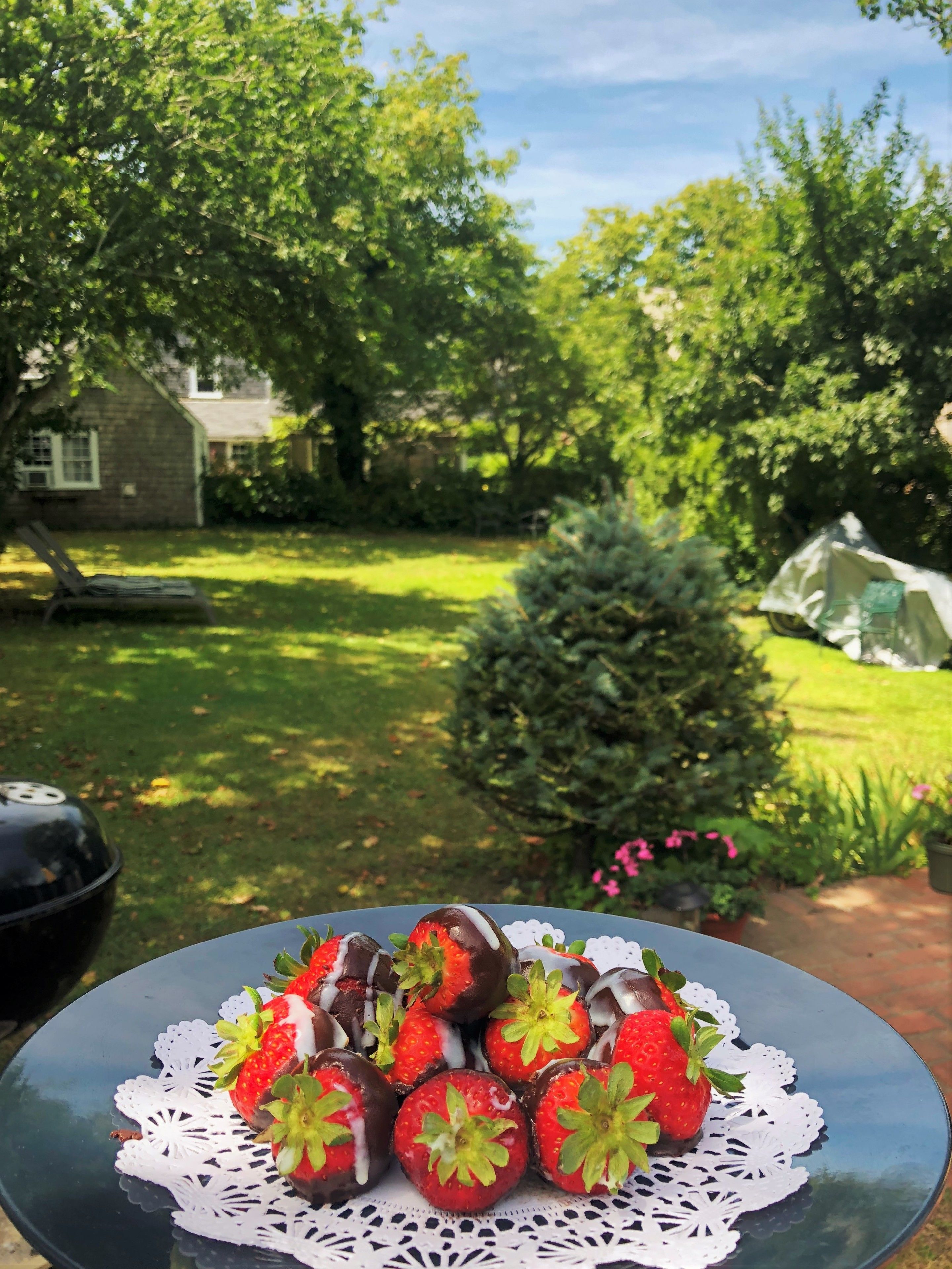 Una mesa con un plato de fresas bañadas en chocolate, ubicada al aire libre con un jardín y una casa al fondo.