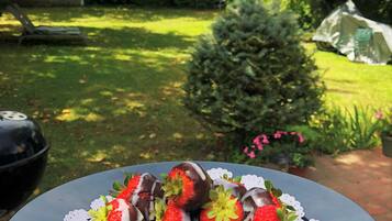 Una mesa con un plato de fresas bañadas en chocolate, ubicada al aire libre con un jardín y una casa al fondo.
