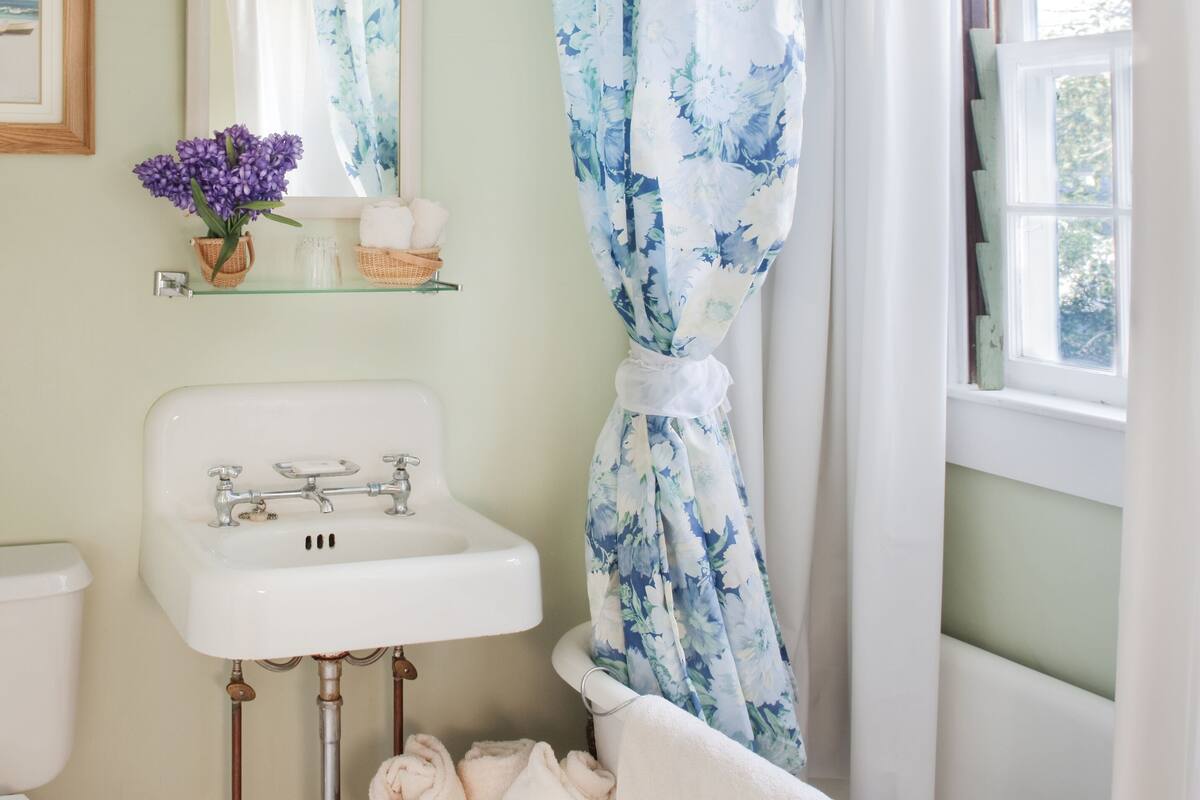 A bathroom with a white sink, a bathtub, a wicker basket with towels, a mirror with a light, and a window with a view.