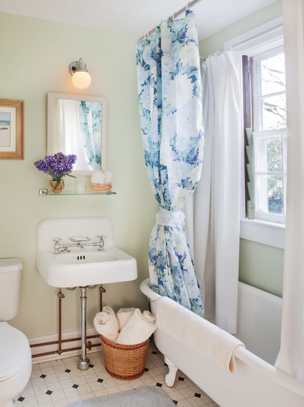 A bathroom with a white sink, a bathtub, a wicker basket with towels, a mirror with a light, and a window with a view.
