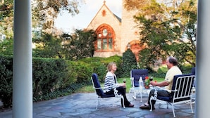 Two people seated at a table with drinks, in a courtyard with stone flooring and flower beds, in front of a church with a stone facade and arched windows.