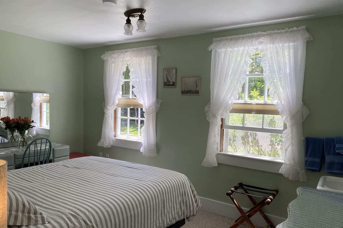 A bedroom with a bed, a dresser, a chair, and two windows with white curtains.