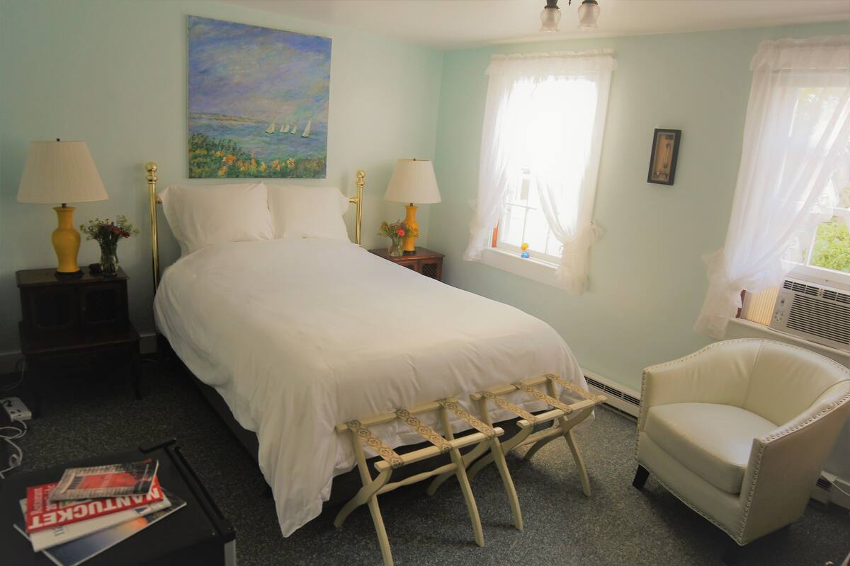 A bedroom with a bed, two bedside tables with lamps, a chair, a window with curtains, and a painting on the wall.