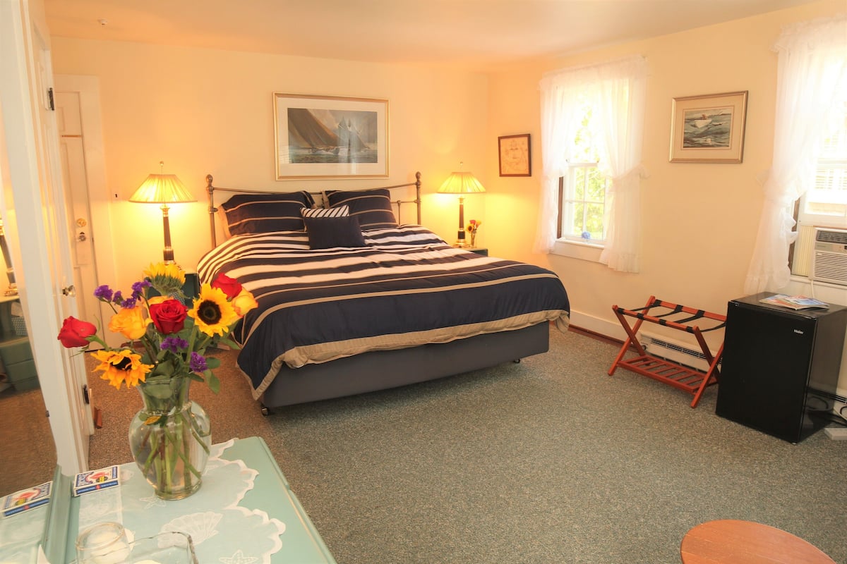 A bedroom with a bed, two bedside lamps, a window with curtains, a radiator, a small table with a vase of flowers, and a picture on the wall.