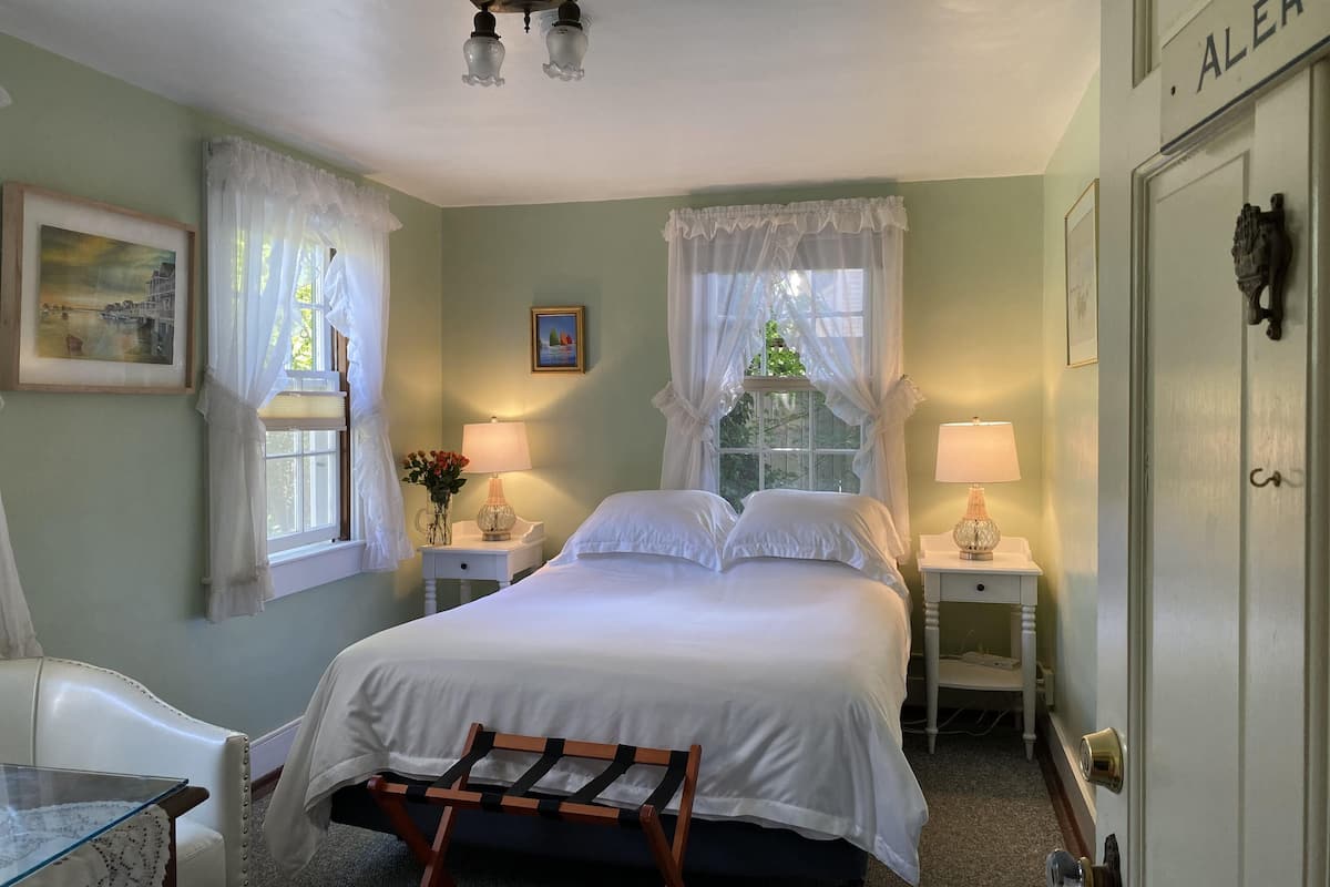 A bedroom with a bed, two bedside tables with lamps, a chair, a bench, and a door with an 'ALERT' sign.