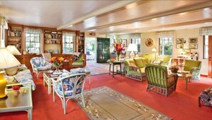 A spacious living room with a red carpet, multiple seating areas with chairs and sofas, a bookshelf, and a door leading outside.
