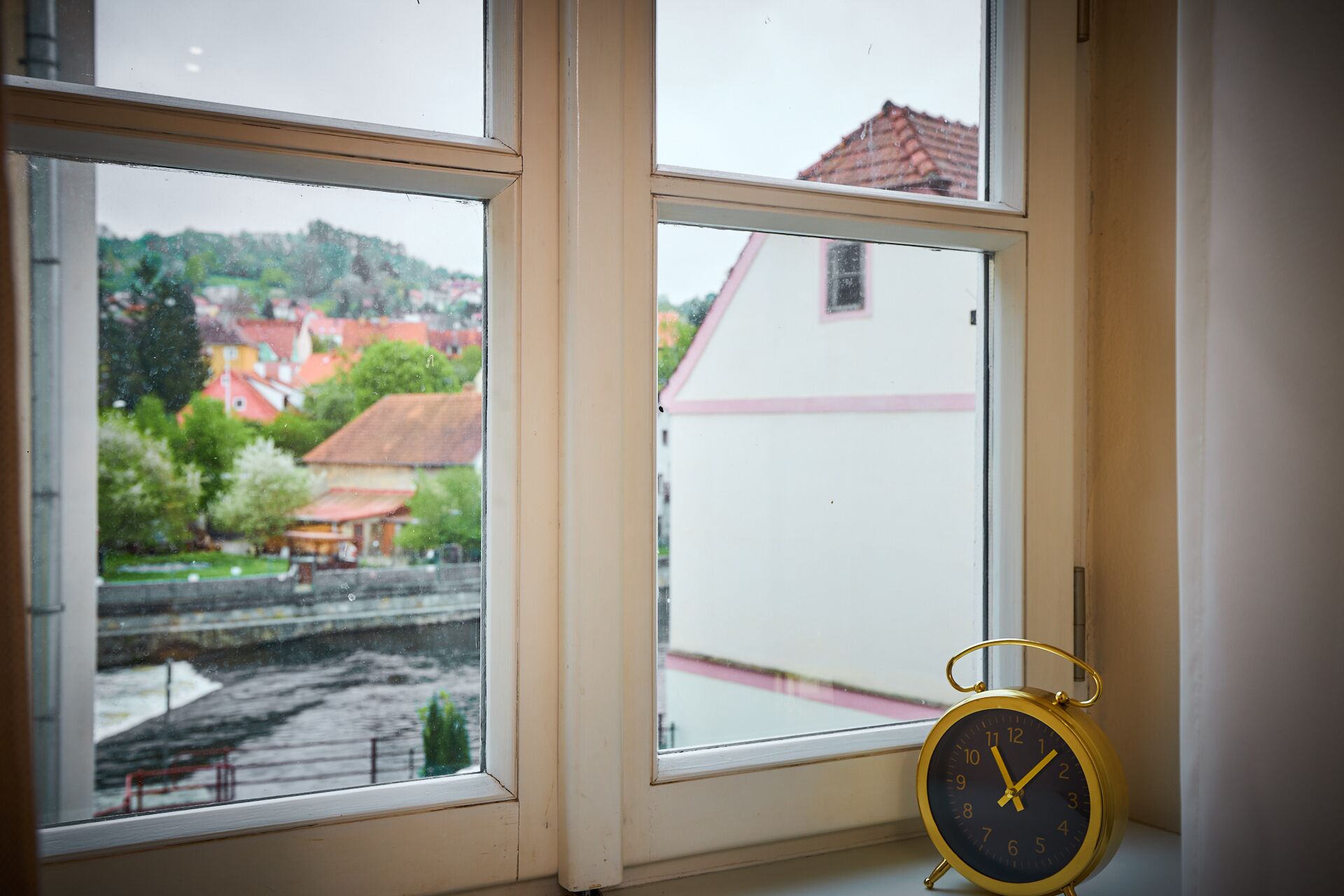 Una ventana con vista a un pueblo, una pared blanca y un despertador dorado.