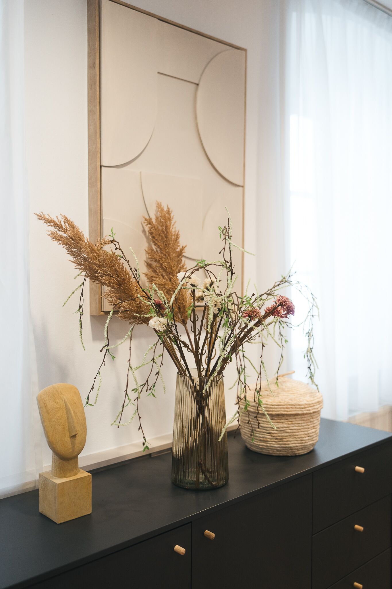 Een modern interieur met een zwarte kast, een houten sculptuur, een glazen vaas met gedroogde planten en een lijst met abstracte kunst.